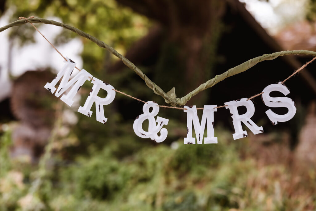 A decorative sign with white letters spelling “MR & MRS” hangs on a string outdoors, with greenery and blurred trees in the background.