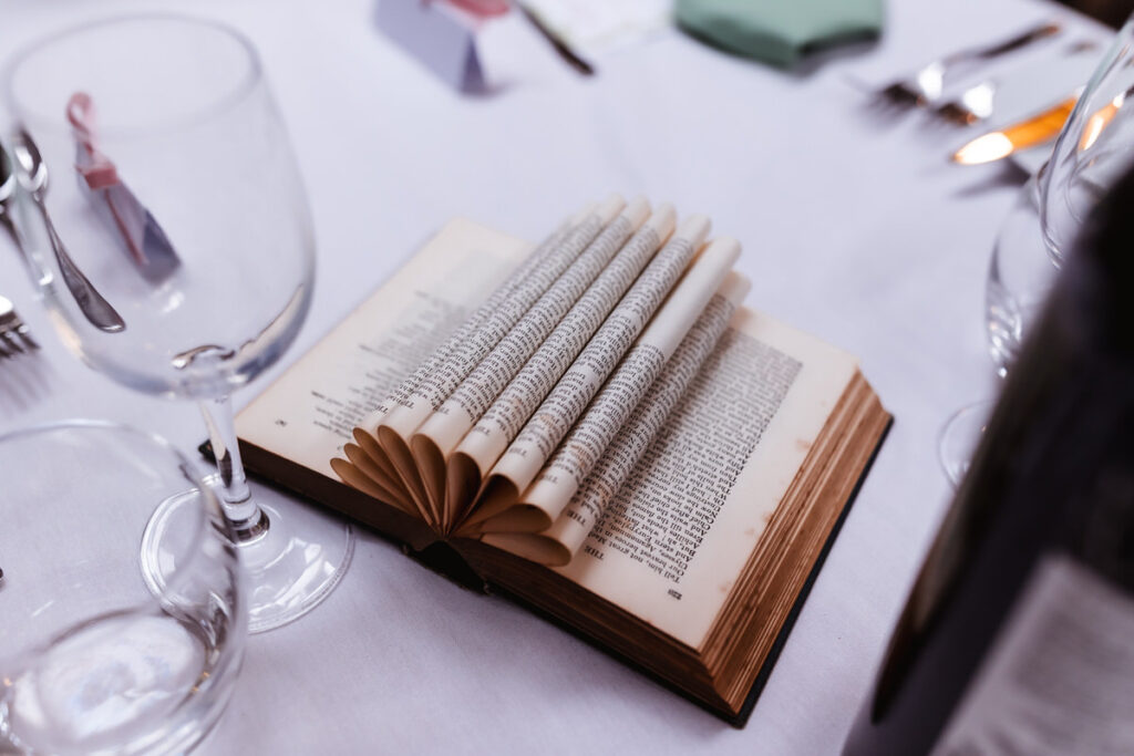 An open book with its pages artistically folded into a fan shape sits on a white tablecloth, surrounded by empty wine glasses, cutlery, and blurred table settings.