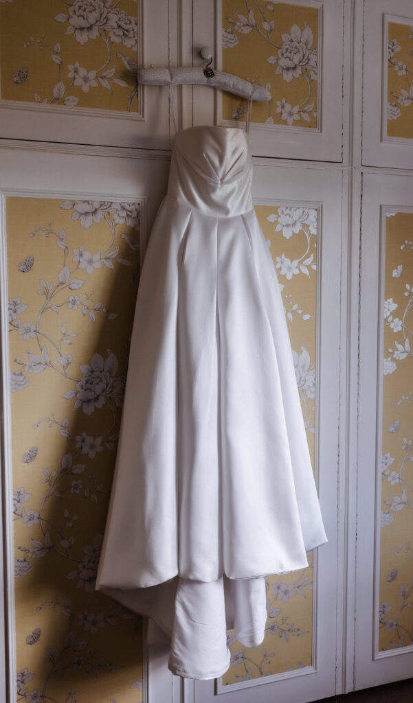 A strapless white wedding dress hangs on a padded hanger against a cream wardrobe with yellow floral-patterned panels. The dress has a fitted bodice and a full, flowing skirt.