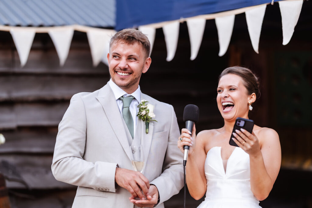 A bride in a white dress laughs while holding a microphone and phone, standing next to a smiling groom in a light suit with a boutonniere, in front of a rustic wooden background and white bunting flags.