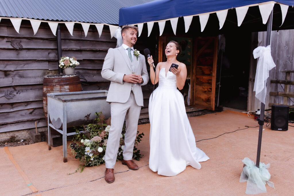 A bride in a white dress and a groom in a light suit stand together outside, smiling and laughing during a speech at their wedding reception, under a canopy decorated with bunting.