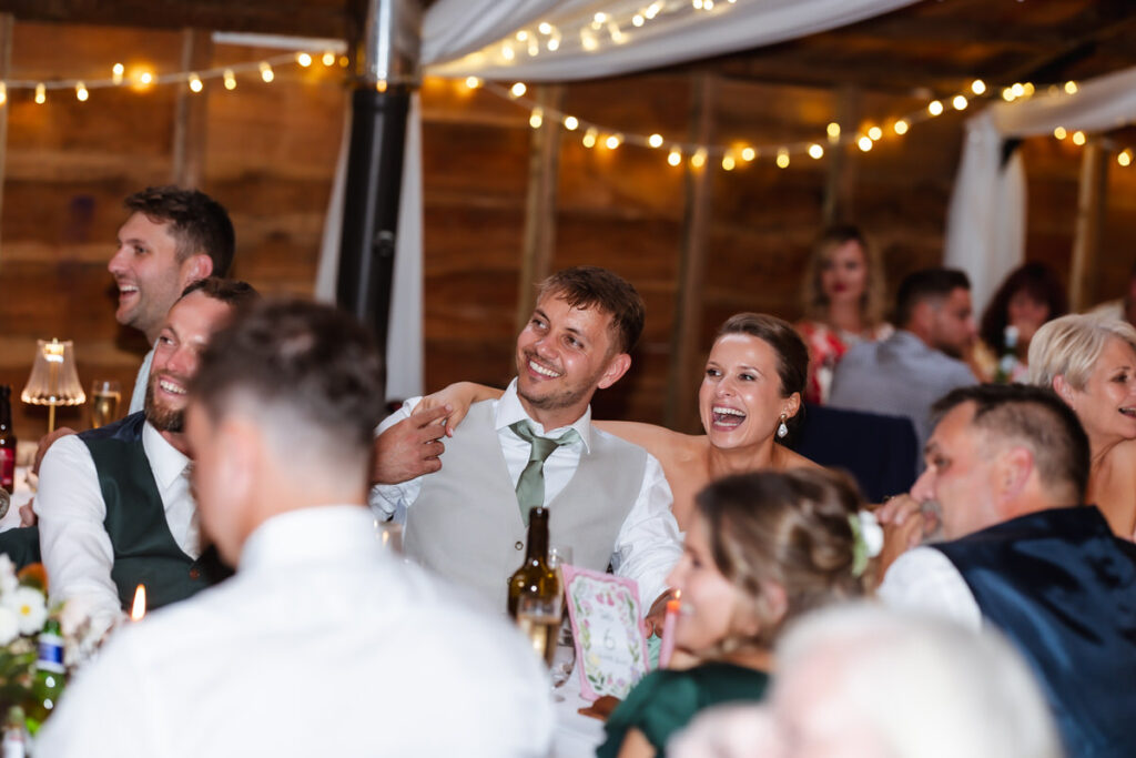 A group of people dressed formally sit smiling and laughing at decorated tables during a celebration inside a warmly lit venue with string lights and wooden walls.