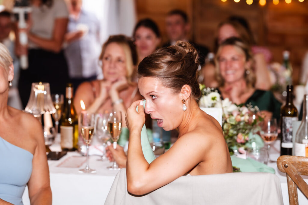 A woman at a formal event wipes away tears with a napkin, looking emotional. She is seated at a table with wine glasses, bottles, and flowers, while others in the background watch and smile.