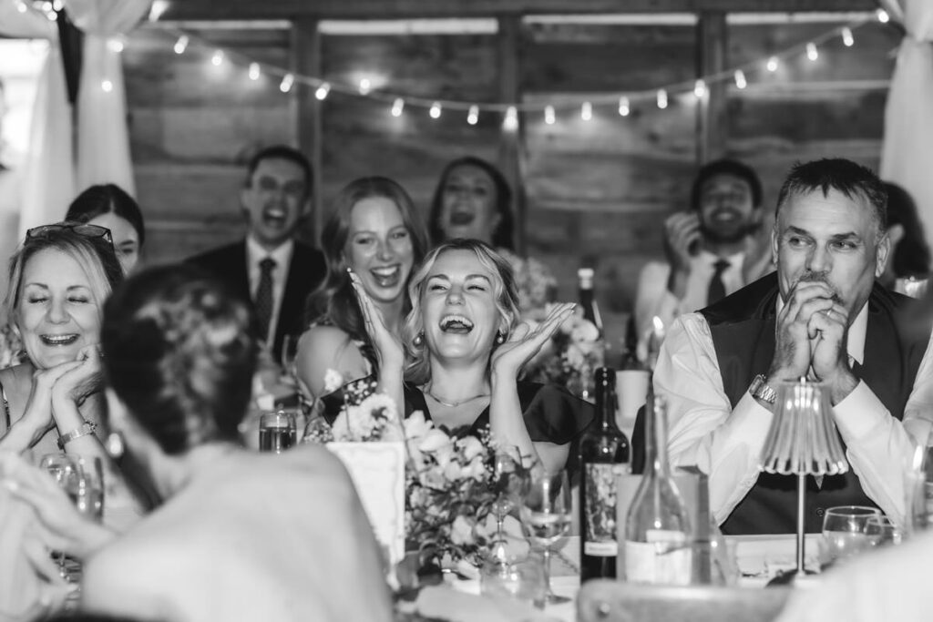 A group of people at a wedding reception sit around a decorated table, laughing and smiling. The focus is on a woman in the center with her hands by her face, surrounded by joyful guests. The image is black and white.