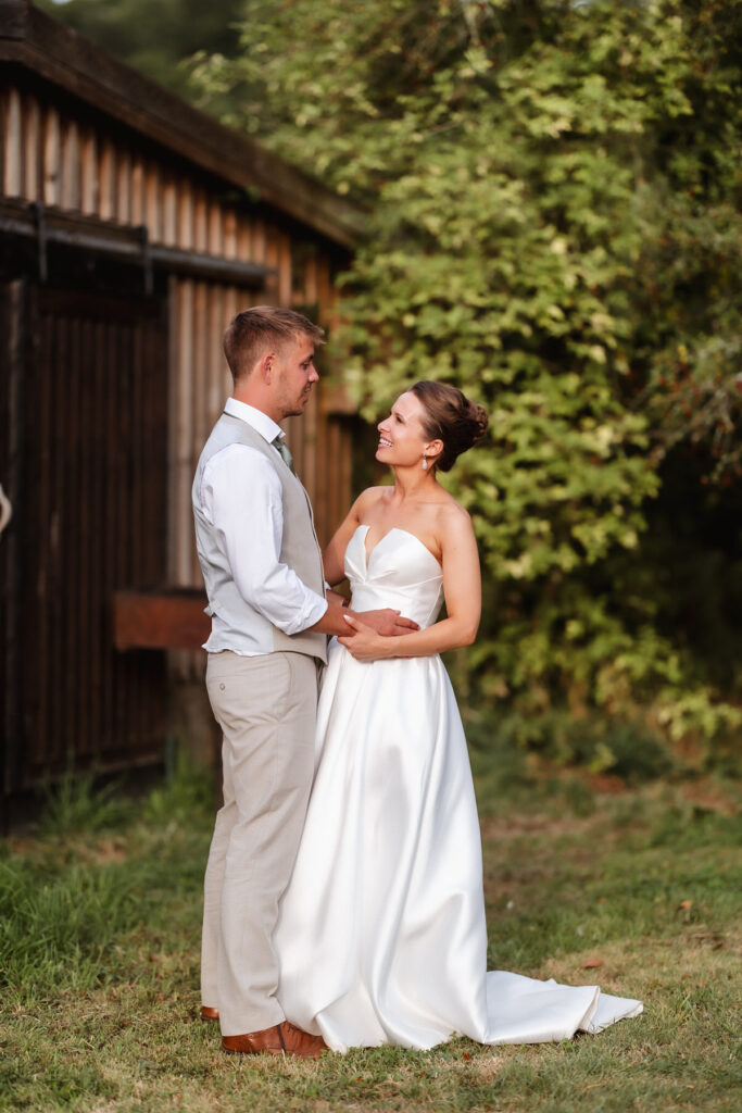 A bride and groom stand outdoors, smiling and holding each other in front of a rustic wooden building and lush greenery. The bride wears a strapless white gown, and the groom is dressed in a light vest and trousers.