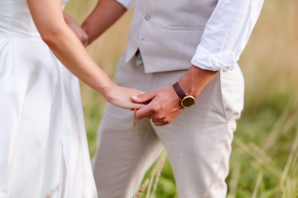 A bride and groom standing outdoors hold hands gently; both are dressed in white, with the groom wearing a light suit and brown watch. The background is grassy and blurred. Their faces are not visible.