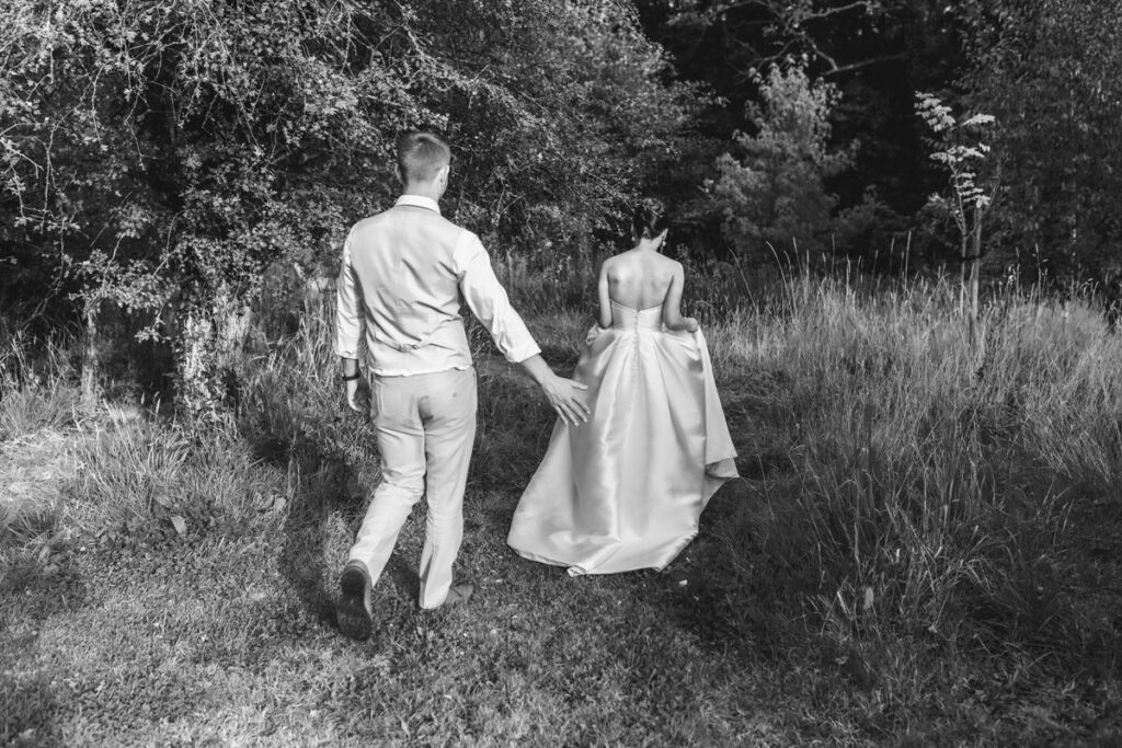 A man and a woman in formal attire, possibly a groom and bride, walk away on a grassy path surrounded by trees. The scene is in black and white, with the woman slightly ahead, holding up her dress.