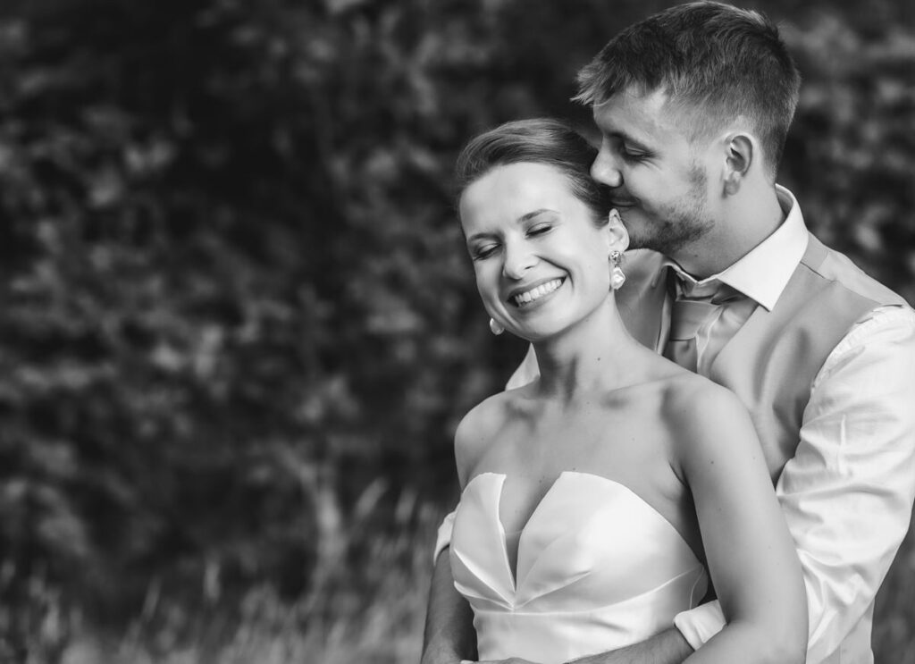 A smiling bride in a strapless gown stands in front of a groom who hugs her from behind. They appear happy and relaxed outdoors, with blurred trees in the background. The photo is in black and white.