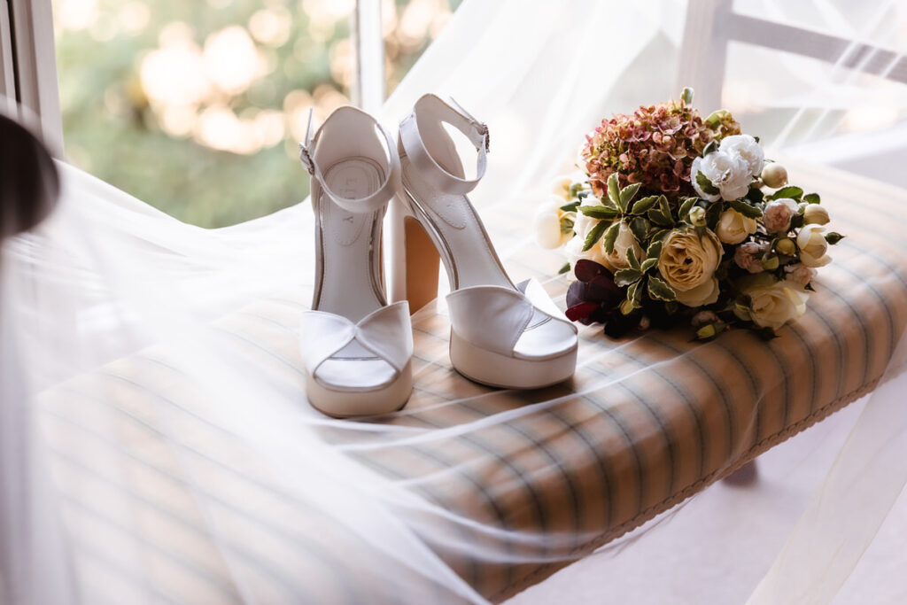 White high-heeled sandals and a bouquet of flowers rest on a striped cushioned bench near a window, with sheer white fabric draped softly around them.