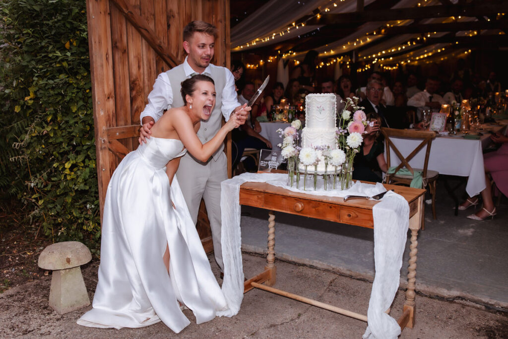 A bride in a white dress excitedly laughs while holding a knife with her groom, preparing to cut their wedding cake. The cake is decorated with flowers and the couple is surrounded by wedding guests in a warmly lit venue.