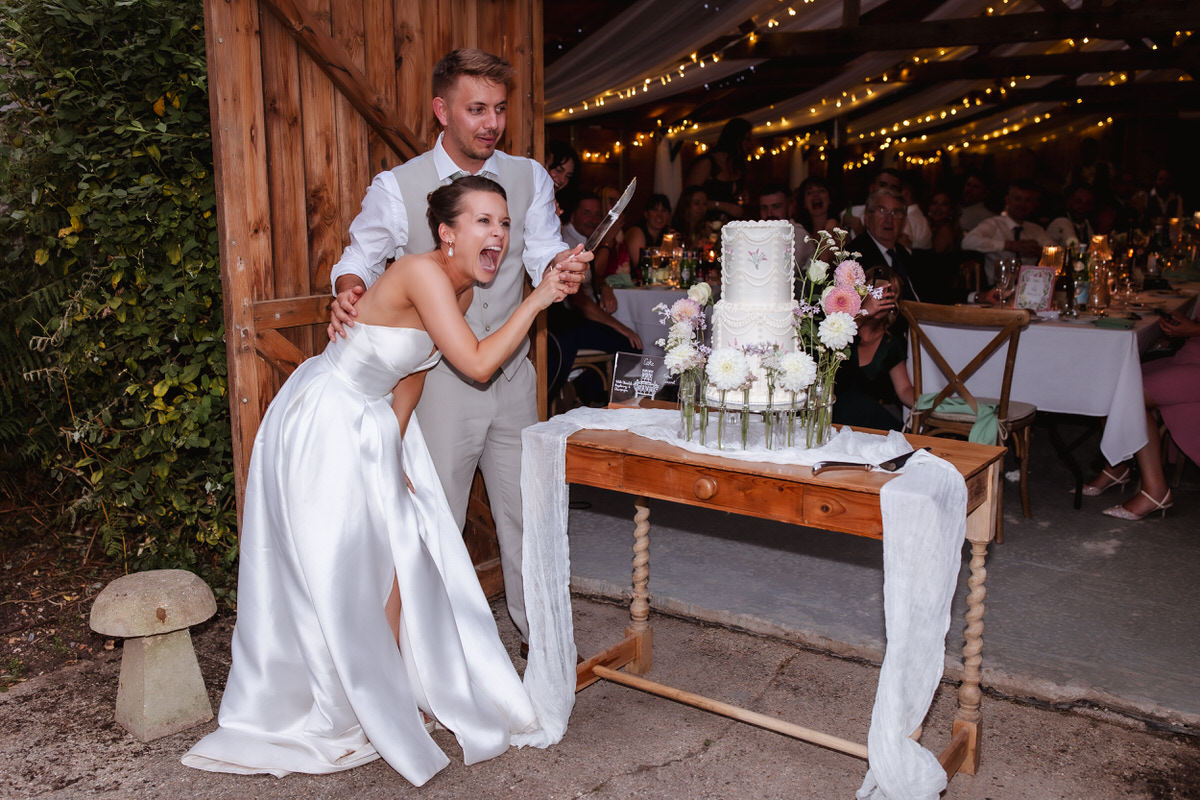 A bride in a white dress excitedly laughs while holding a knife with her groom, preparing to cut their wedding cake. The cake is decorated with flowers and the couple is surrounded by wedding guests in a warmly lit venue.