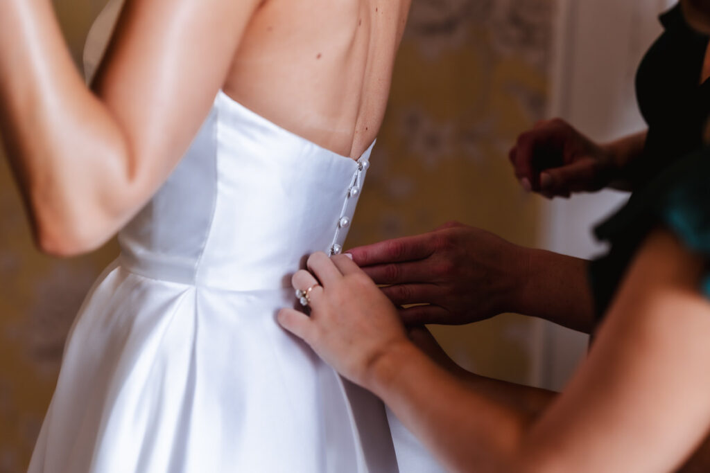 Close-up of someone fastening the buttons on the back of a bride’s white satin wedding dress, with another person assisting. The scene is indoors, and the focus is on the dress and hands.