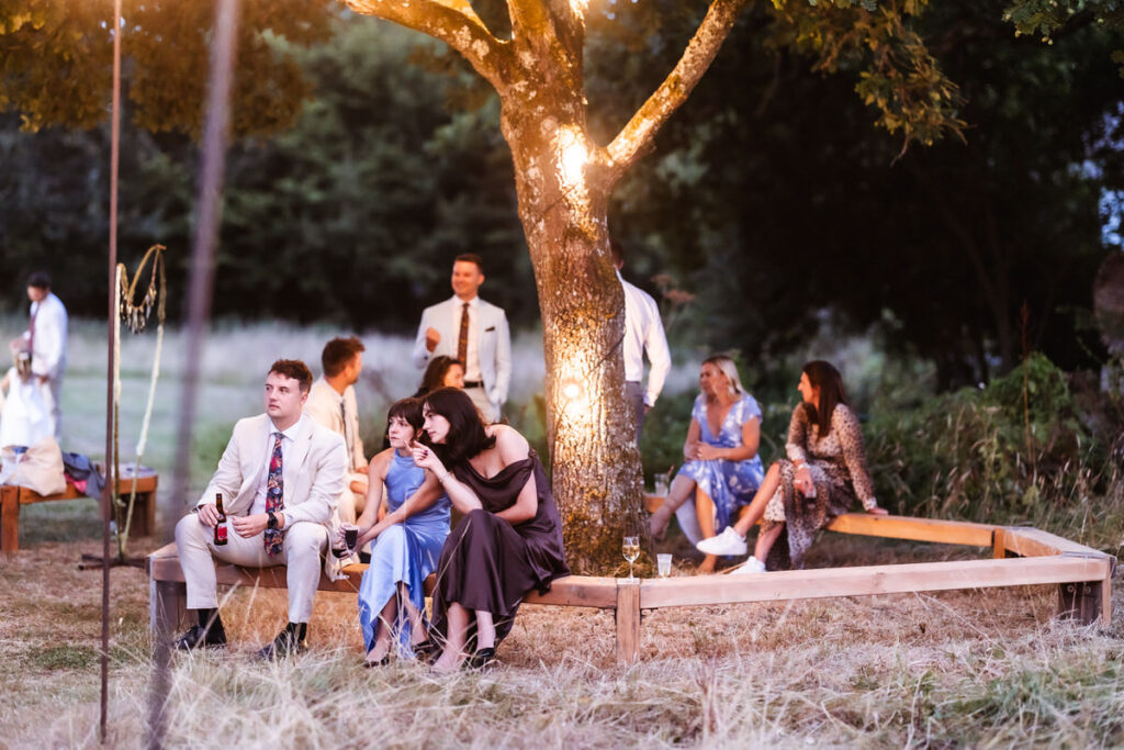 People dressed for a formal outdoor event sit on wooden benches under a tree with string lights, talking and relaxing in a grassy, natural setting. Some people are in the background, while others sit closer to the camera.