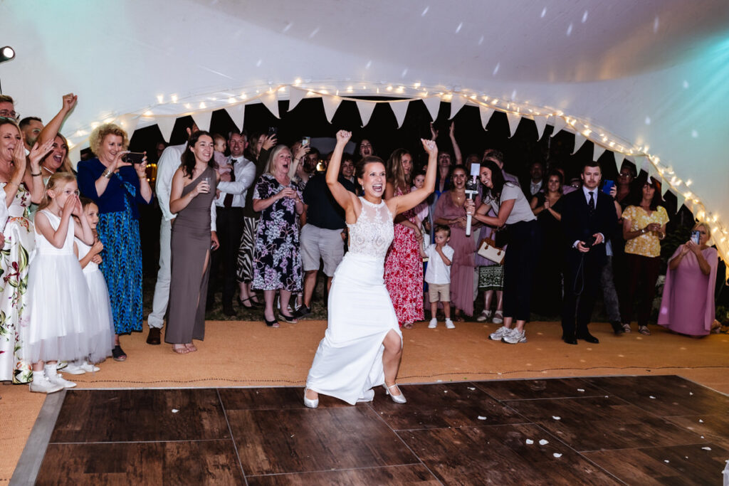 A bride in a white dress dances energetically with arms raised, surrounded by smiling and cheering wedding guests under a tent decorated with string lights.