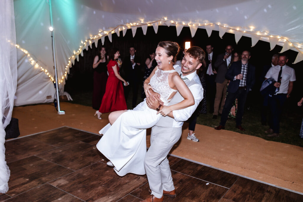 A bride and groom laugh joyfully while dancing under a tent with string lights at night, as guests in formal attire cheer and watch in the background.