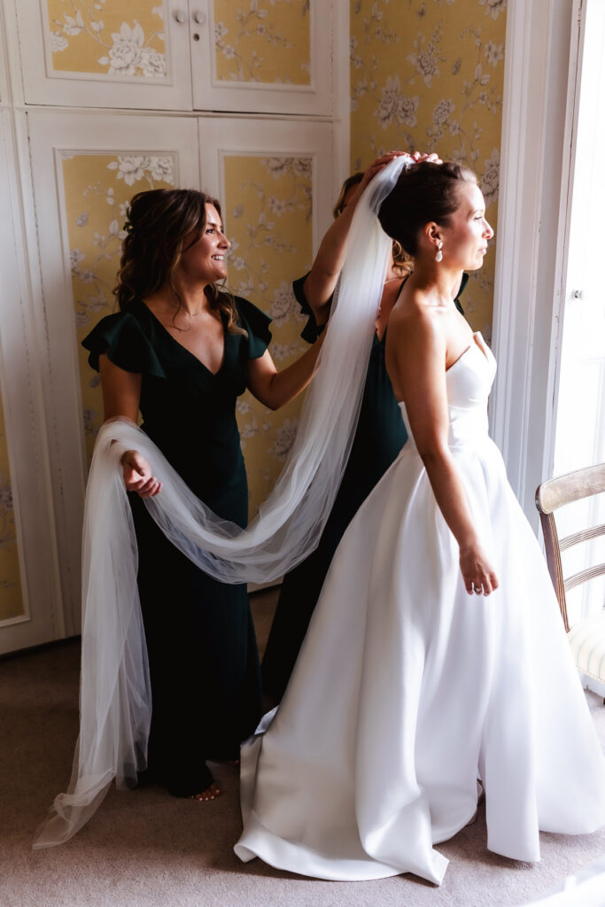Two women in dark dresses help adjust the veil of a bride in a white gown who stands by a window, bathed in natural light, in a room with floral wallpaper and white cabinets.