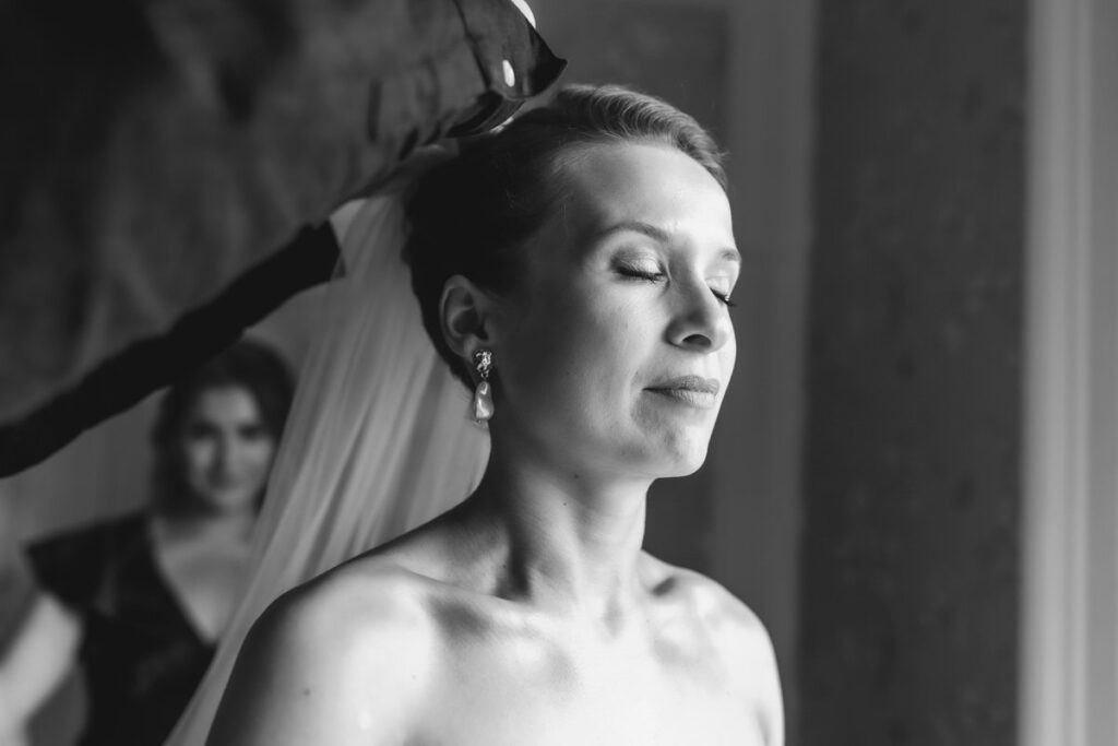 A bride with closed eyes calmly stands as someone adjusts her veil. Another woman, slightly blurred, is visible in the background. The image is in black and white.