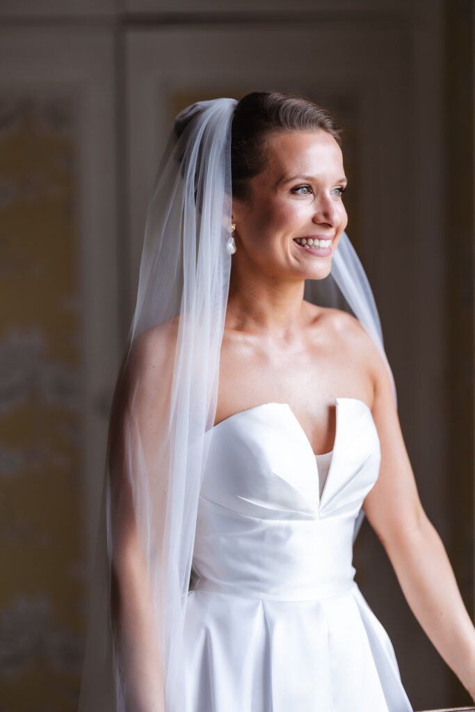 A smiling bride wearing a strapless white wedding dress and a long veil stands indoors, looking to the side with soft natural light illuminating her face.