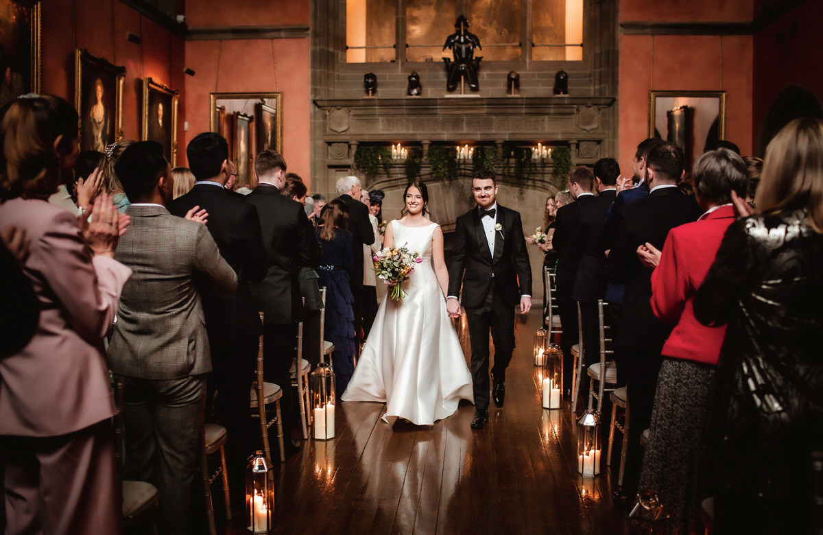 A bride and groom walk down the aisle together in a warmly lit, elegant hall as guests applaud. Captured by a Hampshire wedding photographer, the scene glows with romance—candles line the aisle as love fills the air.