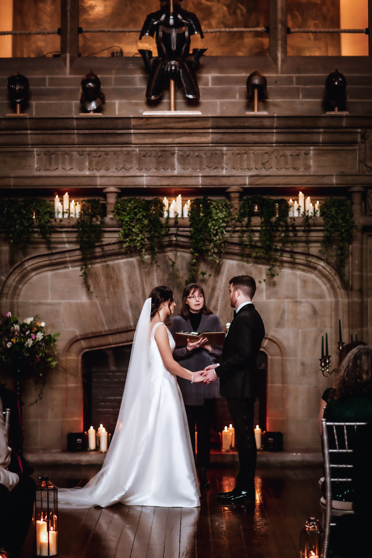A bride and groom stand holding hands during their wedding ceremony in front of a stone fireplace, surrounded by candles and greenery, while a Hampshire wedding photographer captures the beautiful moment.