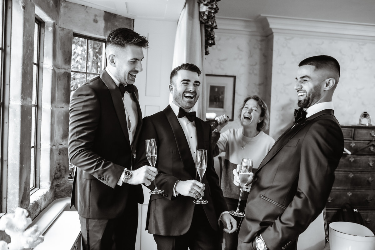 Three men in tuxedos laugh and hold champagne glasses near a window, while a woman joins in the laughter. Captured by a Hampshire wedding photographer, the joyful mood fills this well-lit, celebratory scene.