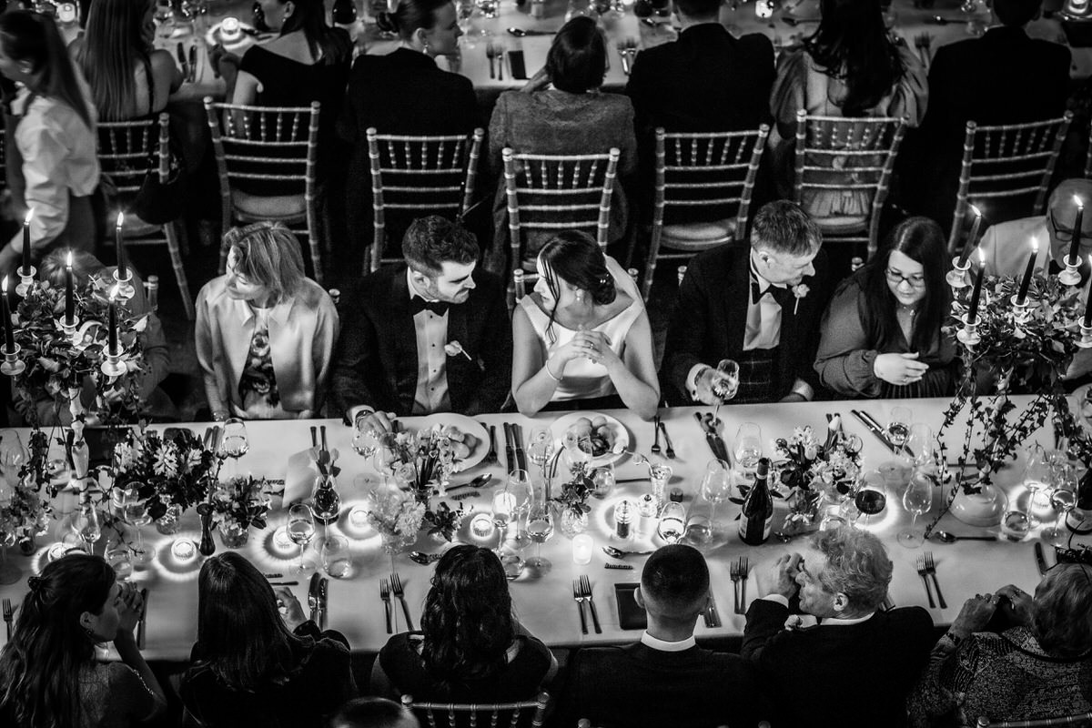 A black and white photo by a Hampshire wedding photographer shows people in formal attire seated at a long, elegant dinner table, decorated with candles and flowers, engaged in conversation and enjoying a festive, celebratory event.