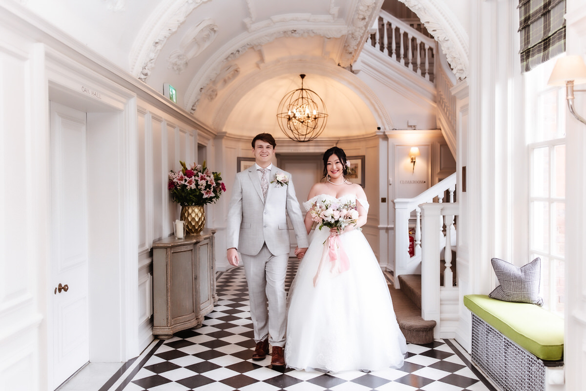 A bride in a white gown and a groom in a light grey suit walk hand in hand along a checkered hallway with white walls and ornate décor, both smiling and carrying a bouquet of flowers, perfectly captured by a Hampshire wedding photographer.