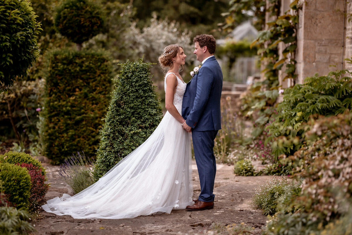 A bride in a flowing white dress and a groom in a blue suit stand holding hands, surrounded by lush greenery and stone walls—captured beautifully by a Hampshire wedding photographer.