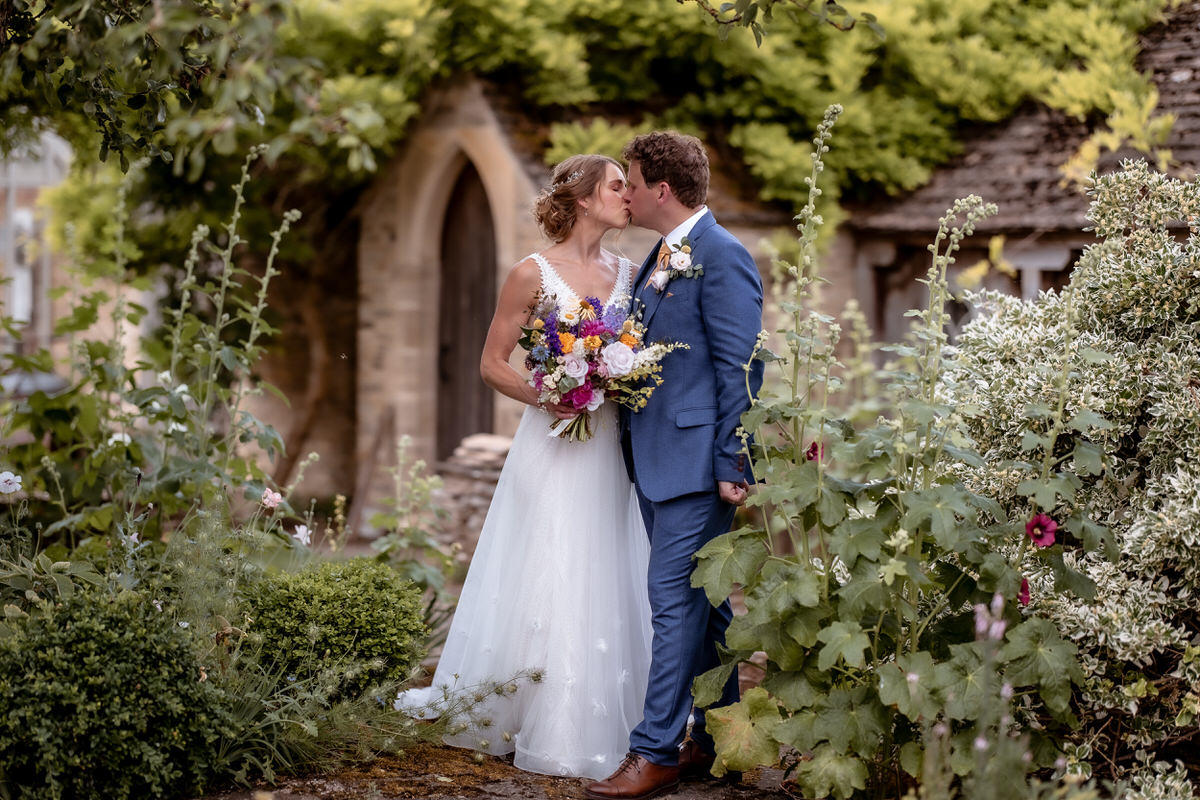 A bride and groom share a kiss in a lush garden, surrounded by greenery and flowers. Captured by a Hampshire wedding photographer, the bride holds a colorful bouquet as the groom stands beside her, with a stone building in the background.