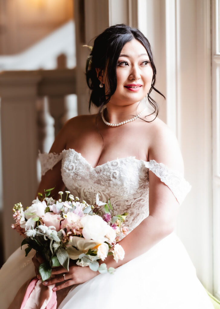 A bride in an off-shoulder white lace wedding gown holds a bouquet and smiles while looking out a bright window, with natural light softly illuminating her face—captured beautifully by a Hampshire wedding photographer.
