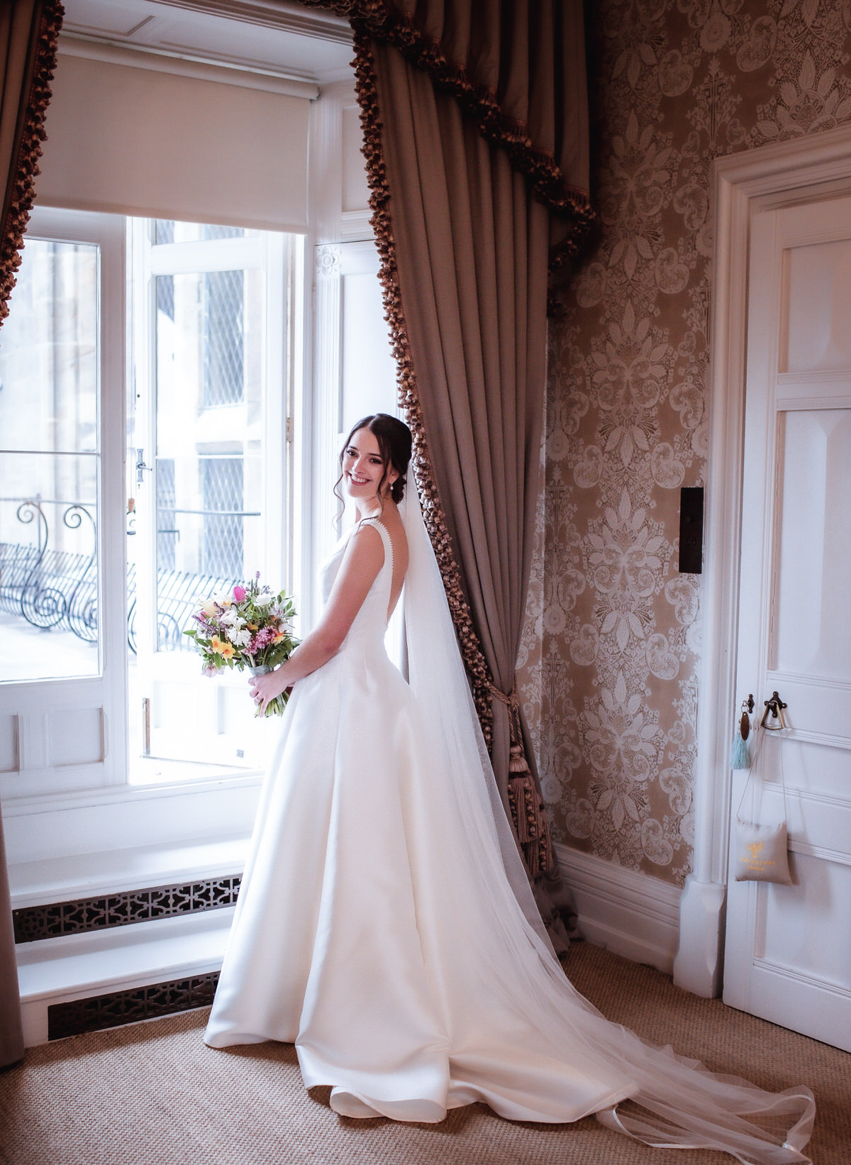 A bride in a white wedding gown and veil smiles by a window, holding a colorful bouquet in a softly lit, elegant room—a timeless moment captured by a Hampshire wedding photographer.