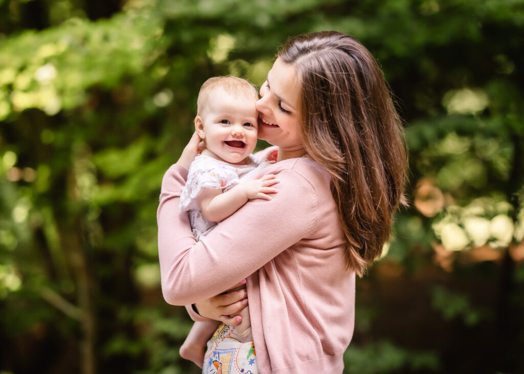 A smiling woman in a pink sweater holds a happy baby outdoors, surrounded by lush green trees and natural light.