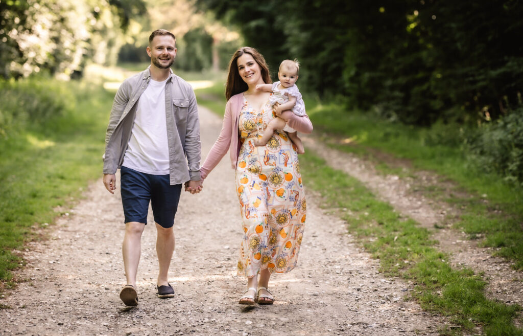 A smiling couple walks hand-in-hand down a sunlit, tree-lined path. The woman carries a baby on her hip, and all three appear relaxed and happy, enjoying a pleasant day outdoors.