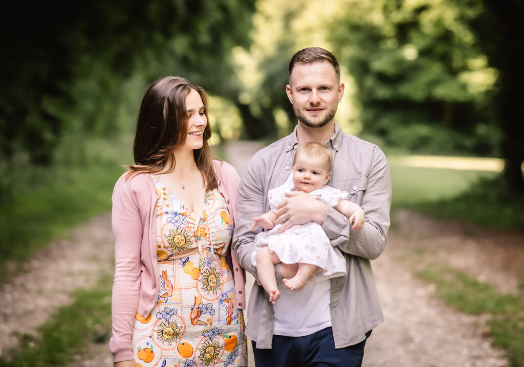 A man and woman stand together on a tree-lined path, smiling, as the man holds a baby in his arms. The woman wears a colorful dress and pink cardigan; the man wears a light shirt. The background is green and leafy.