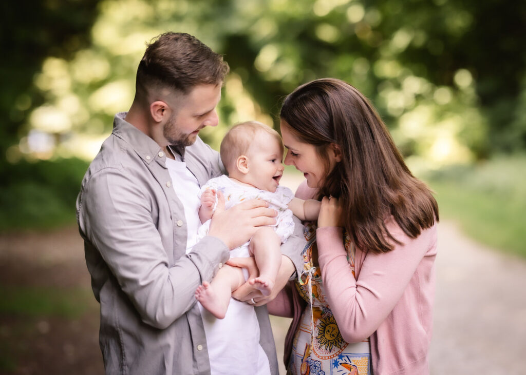 A man and woman smile lovingly at a baby they are holding outdoors, surrounded by green trees and soft sunlight. The baby is reaching towards the woman, and all three appear happy and affectionate.