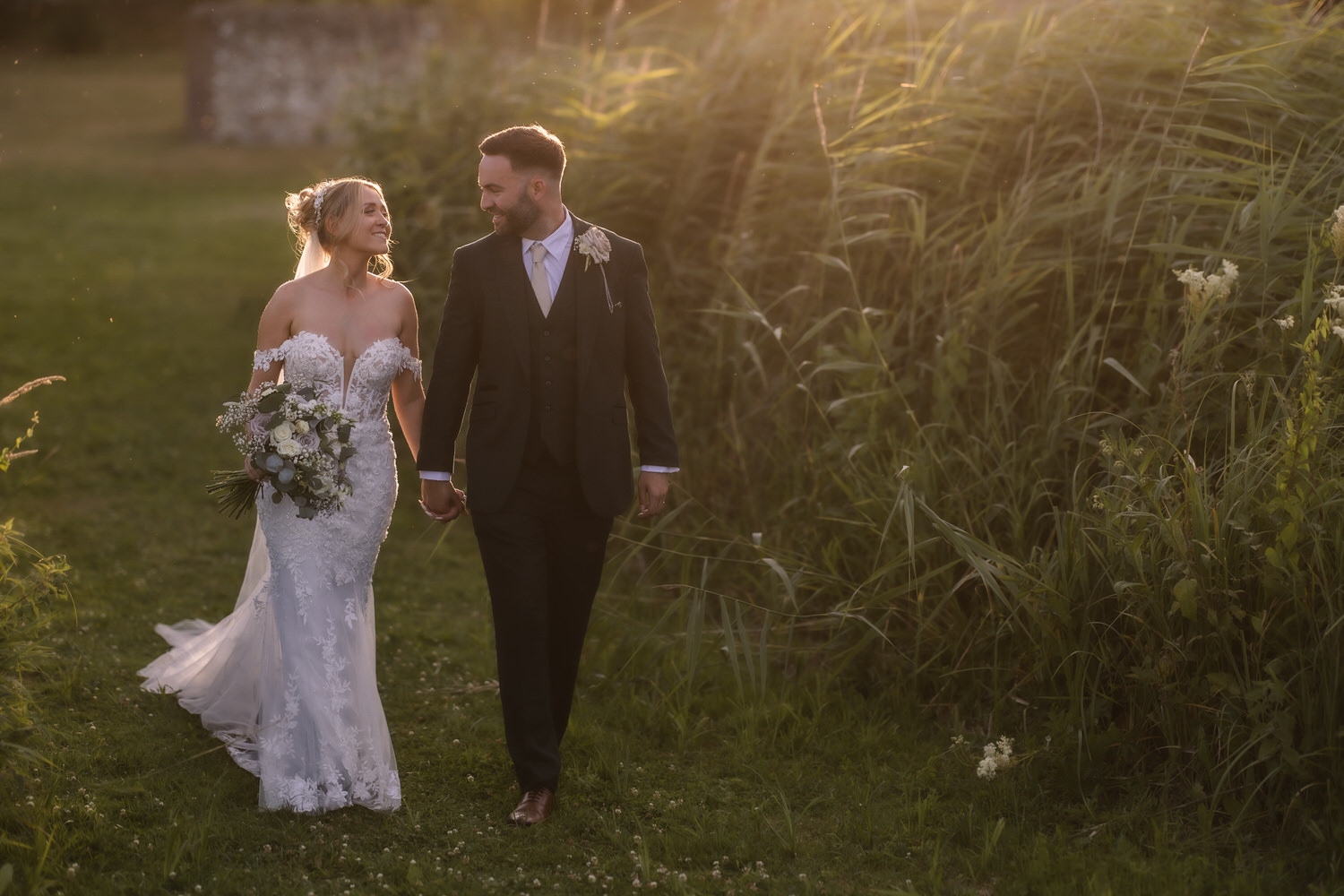 A bride and groom walk hand in hand outside, smiling at each other. Captured by a Hampshire wedding photographer, the bride’s white lace dress and bouquet glow in the golden sunlight as they stroll through tall grass.