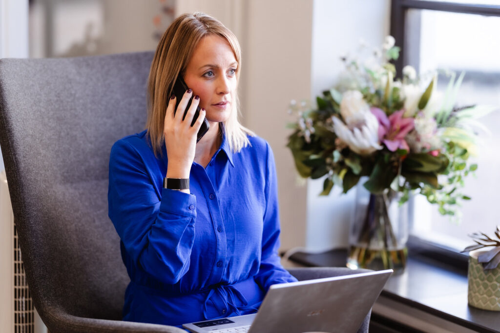 A woman in a blue dress sits in an armchair, holding a phone to her ear and looking thoughtful. She has a laptop on her lap and is next to a window with a bouquet of flowers on the table beside her.