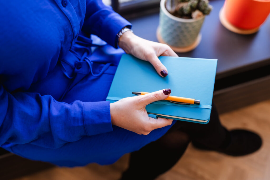 A person in a blue dress is sitting and holding a closed blue notebook with an orange pen on top. There are potted plants in the background on a windowsill.