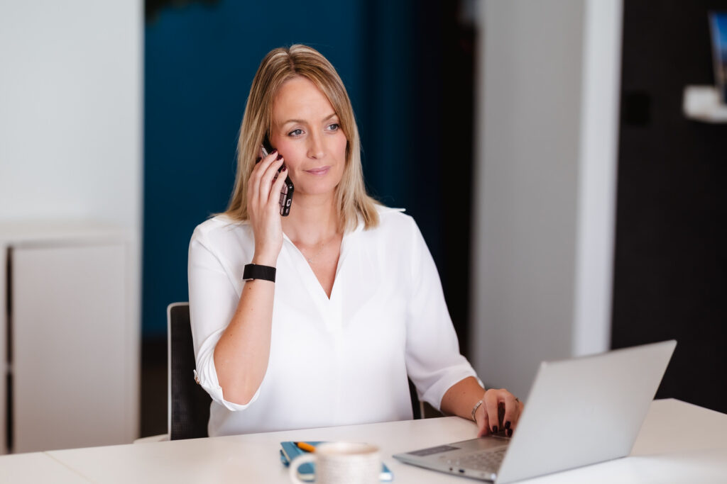 A woman with blonde hair, wearing a white blouse, sits at a desk using a laptop and talking on her phone. A notebook, pen, and coffee cup are on the desk in front of her.