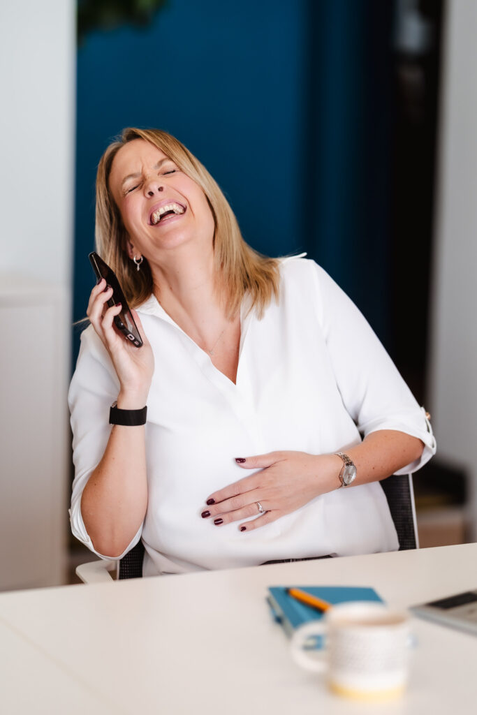 A woman with blonde hair wearing a white blouse sits at a desk, laughing joyfully while talking on her smartphone. She holds her stomach with one hand and has a notebook, pen, and mug in front of her.