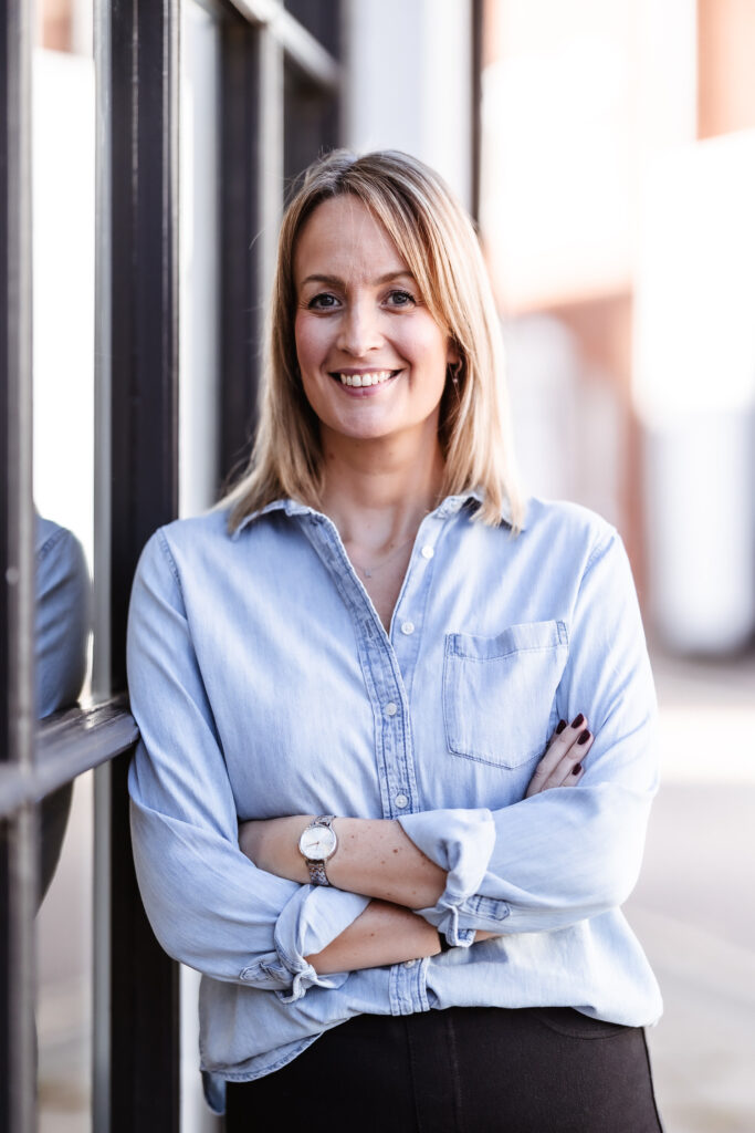 A woman with shoulder-length blonde hair, wearing a light denim shirt and a watch, stands with arms crossed and smiles, leaning against a reflective window. The background is softly blurred.