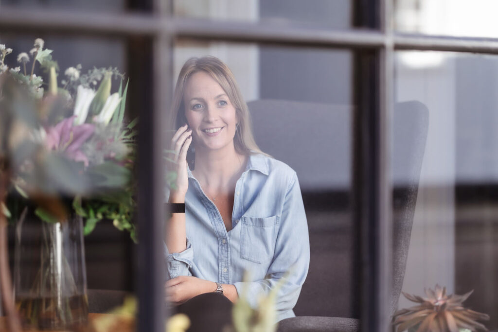 A woman with light brown hair, wearing a light blue button-up shirt, sits in a chair and smiles while talking on the phone. She is seen through a window with reflections of flowers in the foreground.