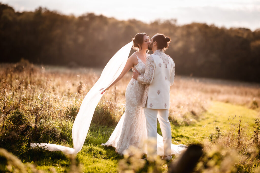 A couple in wedding attire stands in a sunlit field, embracing and smiling at each other. Captured by a Hampshire wedding photographer, the bride’s long veil flows behind her while the groom wears a cream embroidered jacket and white pants.