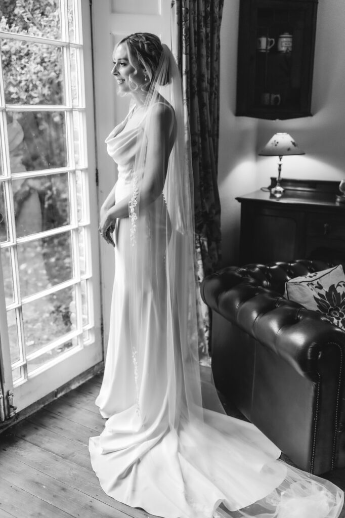 A bride in a long, flowing gown and veil stands by a window, smiling and looking outside. The room is softly lit, with a leather sofa, lamp, and cabinet in the background. The image is in black and white.