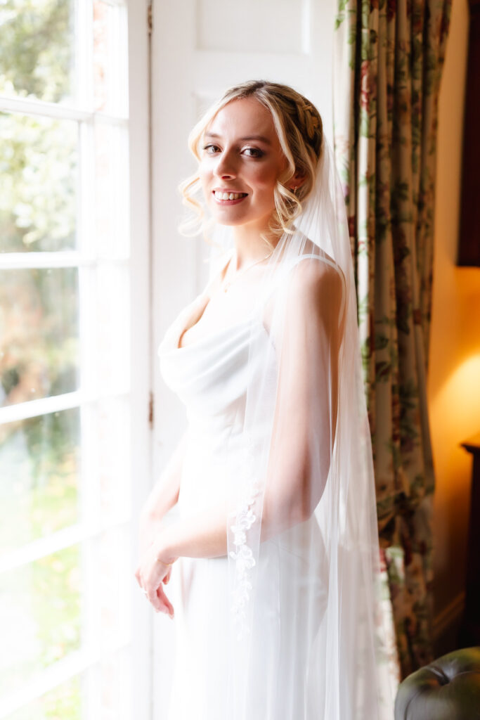 A bride in a white dress and veil stands by a bright window, smiling gently. Light filters through the window, illuminating her face and the delicate details of her dress. Floral curtains hang beside her.