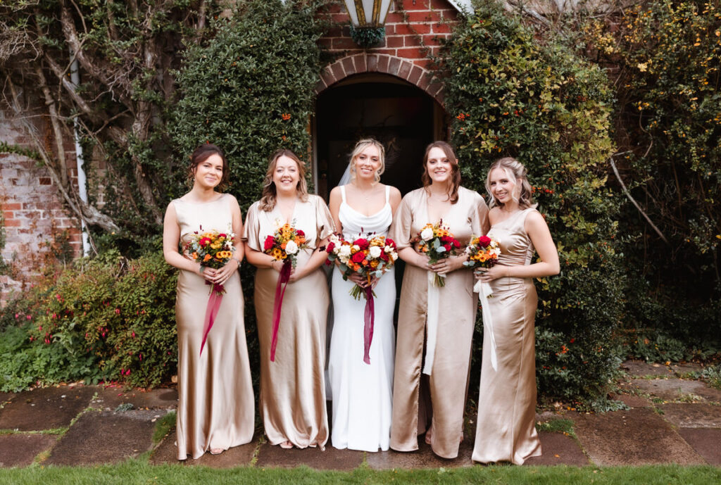 Five women stand side by side outdoors, with the central woman in a white dress and the others in gold dresses, all holding vibrant flower bouquets in front of a leafy brick archway.