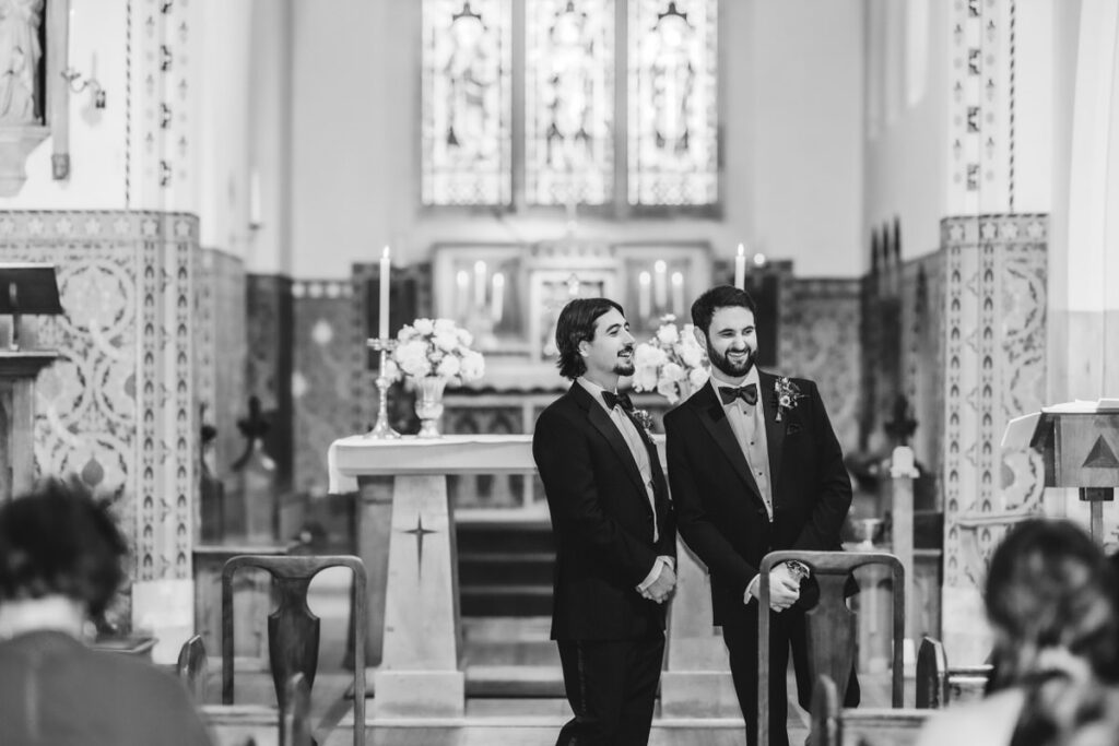 Two men in suits stand smiling at the front of a church, with floral arrangements and lit candles on the altar behind them. The scene is formal and appears to be part of a wedding ceremony.