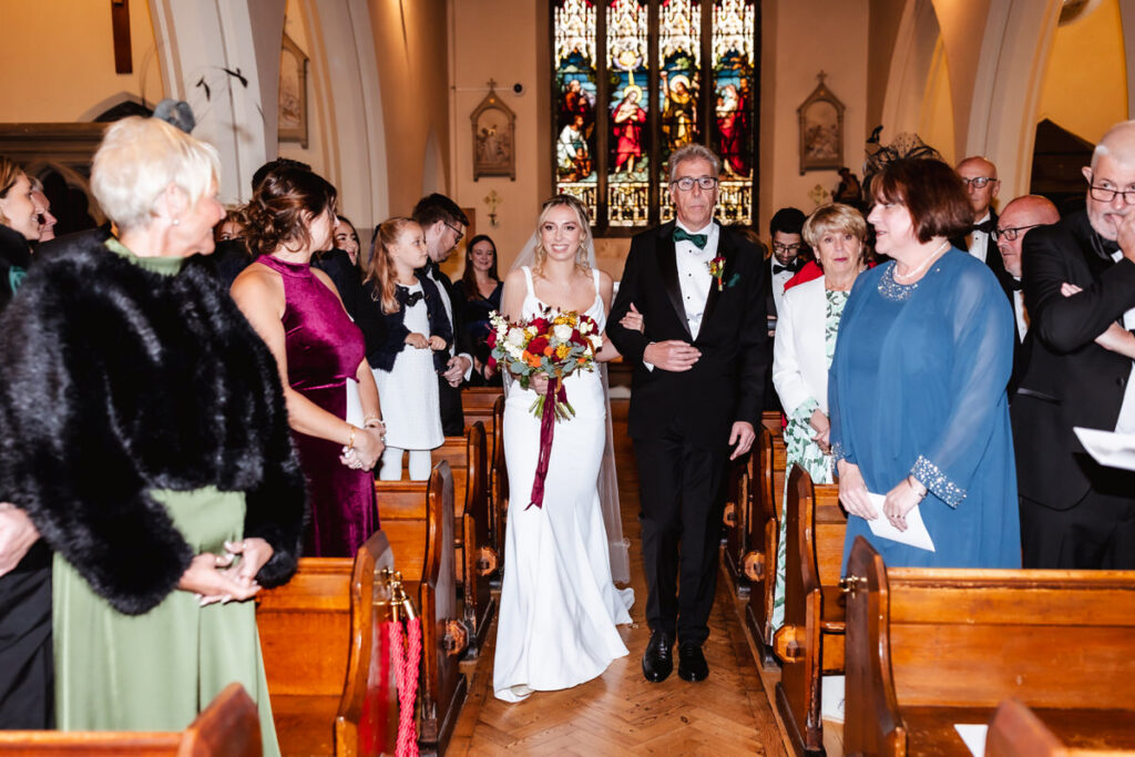 A bride in a white dress and a man in a tuxedo walk down the aisle of a church, surrounded by guests who are seated and standing, with stained glass windows in the background.