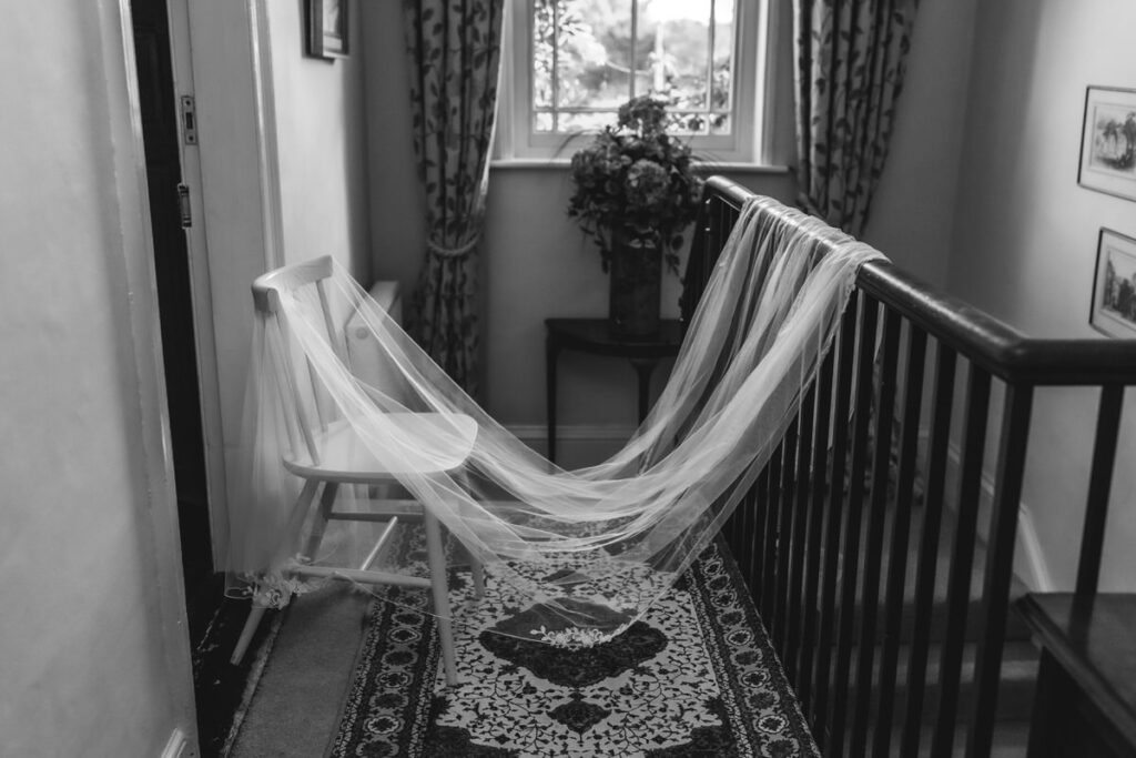 A long bridal veil drapes over a white chair and trails onto a patterned rug in a hallway, with a window, curtains, framed pictures, and a floral arrangement visible in the background.