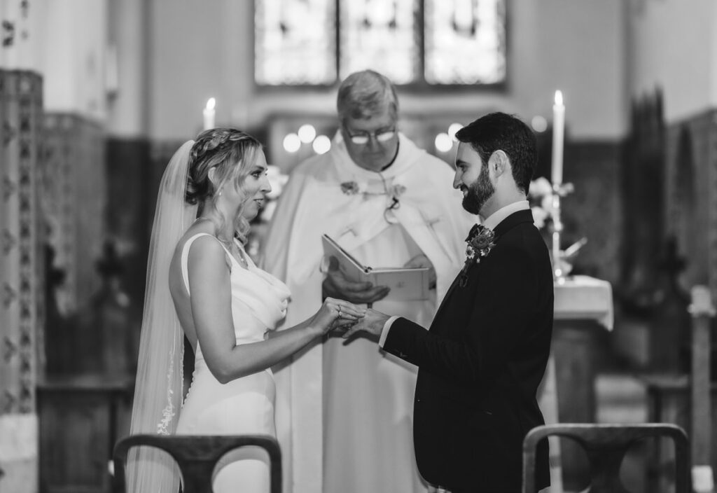 A bride and groom hold hands and exchange vows in a church ceremony, with a priest standing behind them, all dressed formally. The scene is in black and white.